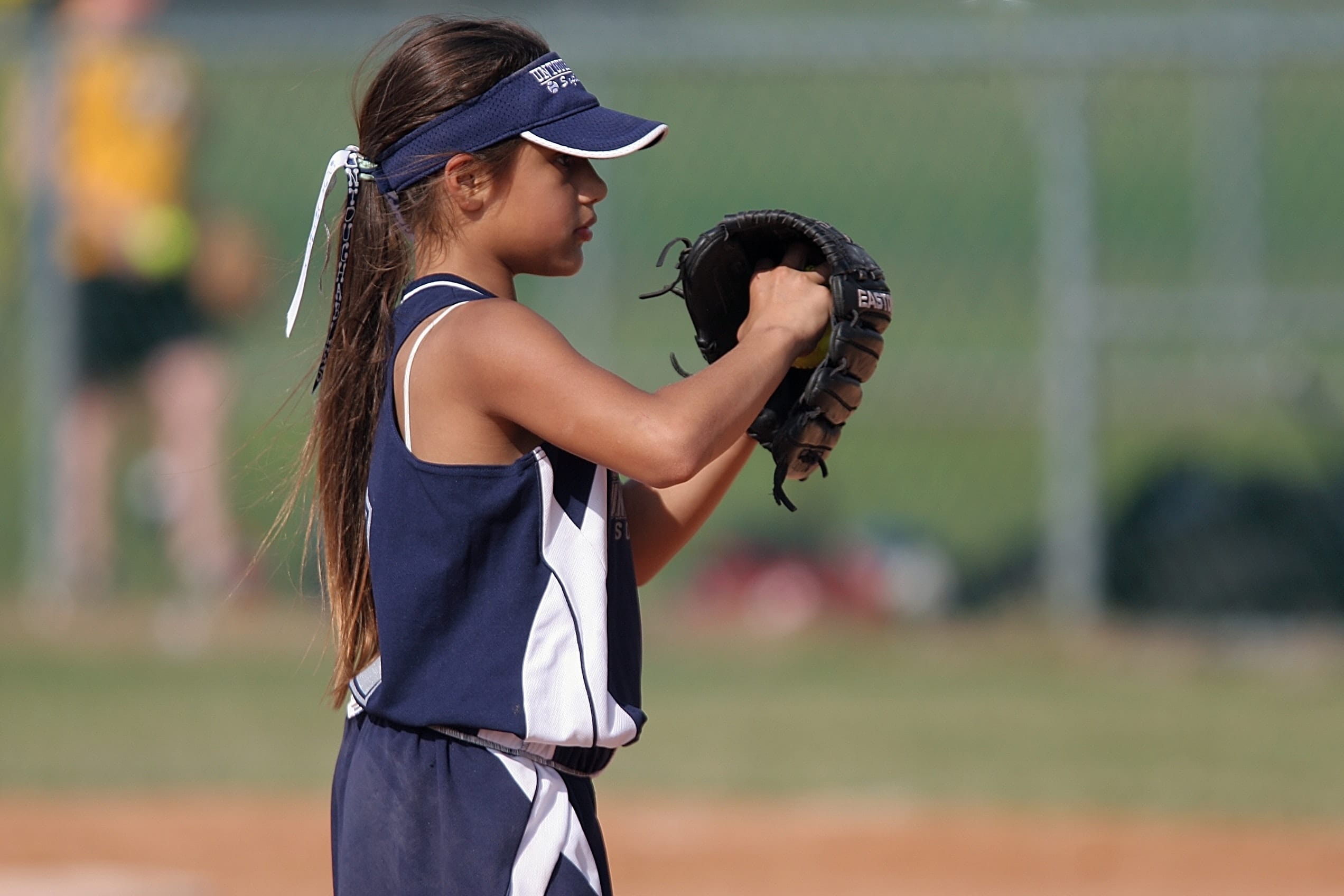 Girl on a baseball diamond in uniform with her right hand in her gloved left hand.